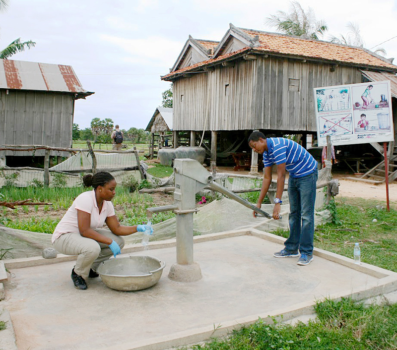 A young woman and a young man at a water pump, pumping water into a bottle and catch basin. Wooden structures are in the background.
