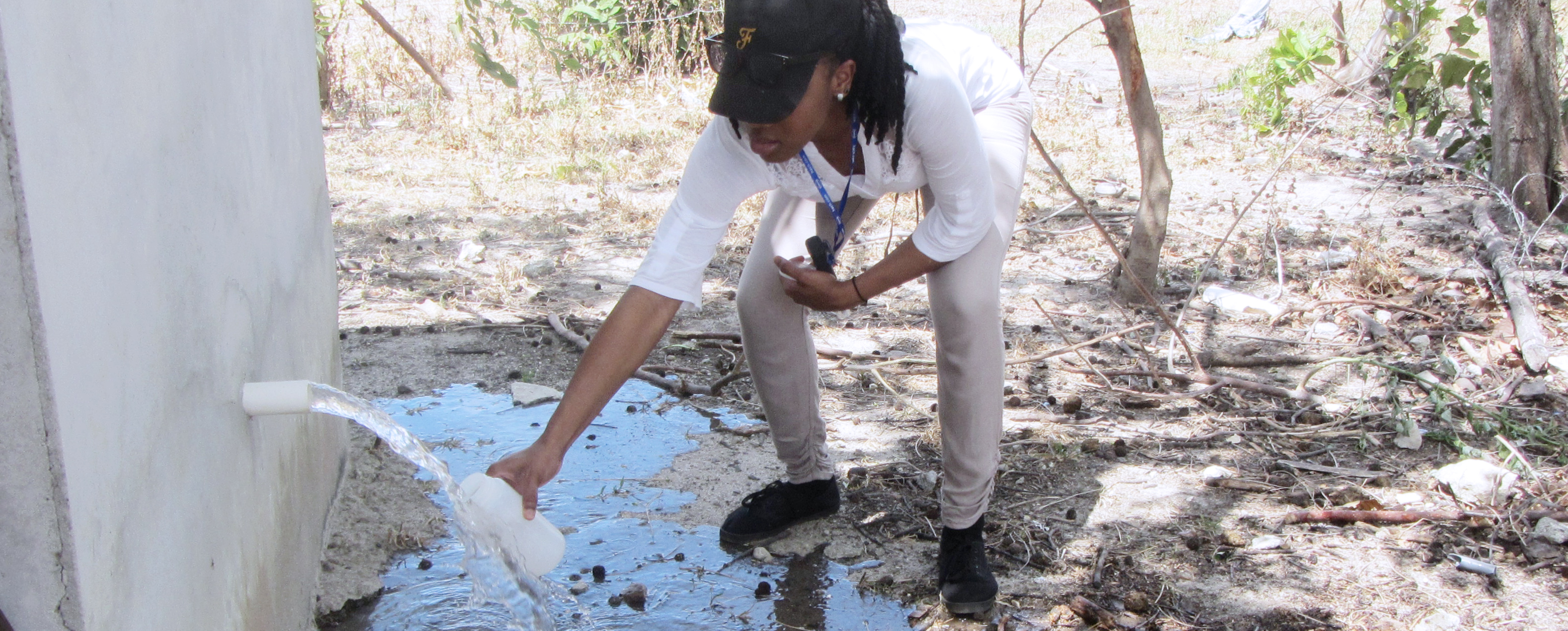 A student in a white shirt and a ball cap is crouching down to fill a water bottle from a spout to perform a water test.