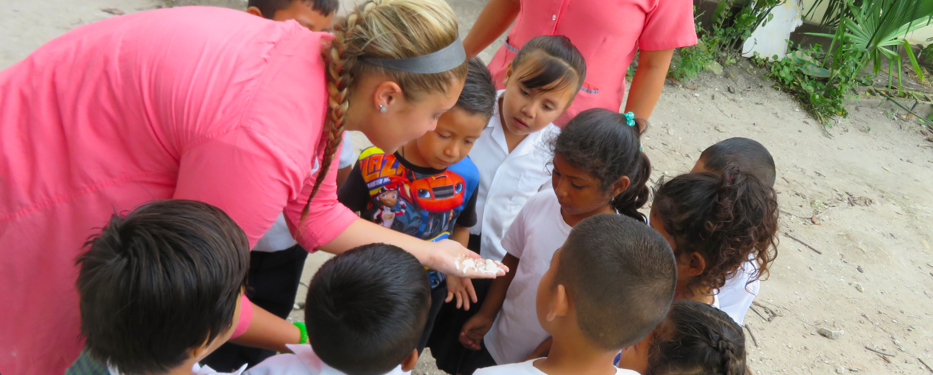 Blonde student surrounded by ten small children who are looking at a light colored substance on her palm which she is holding out.