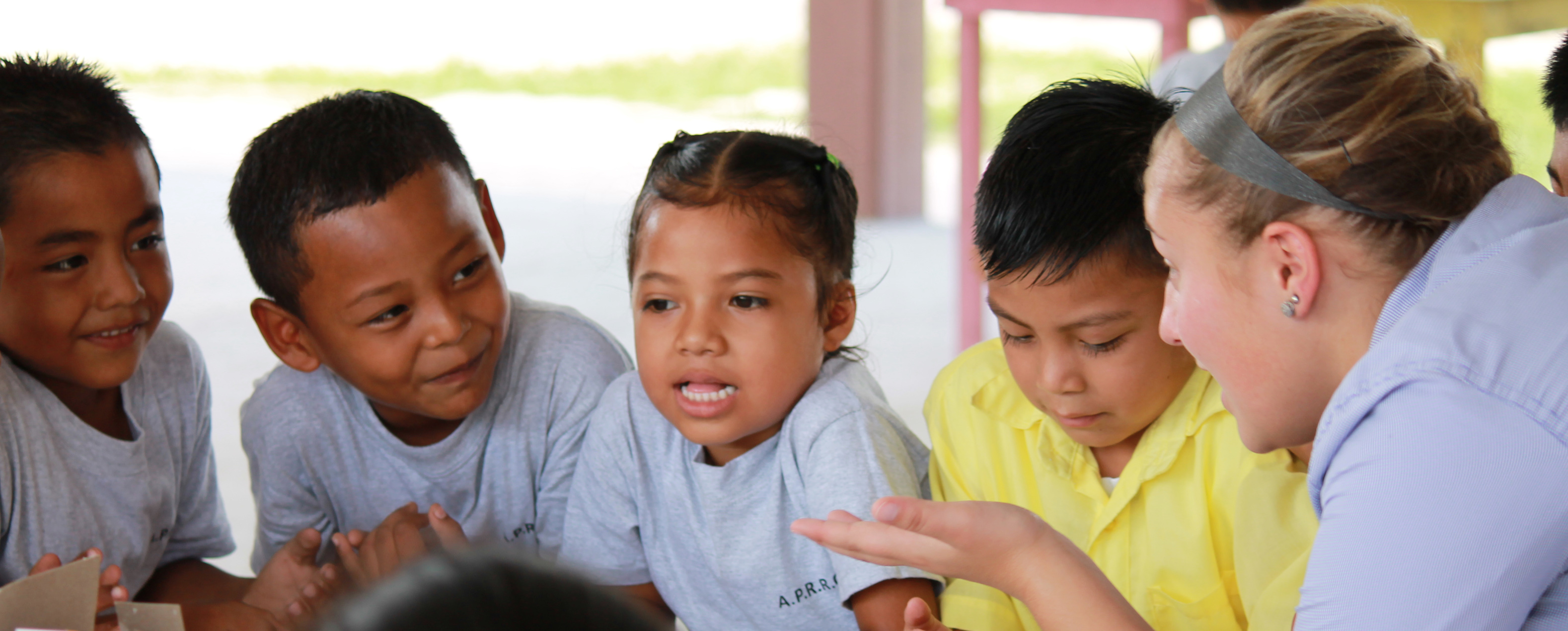 Three small boys and one small girl with black hair gathered around and leaning on a table with an older blonde girl who is talking to them.