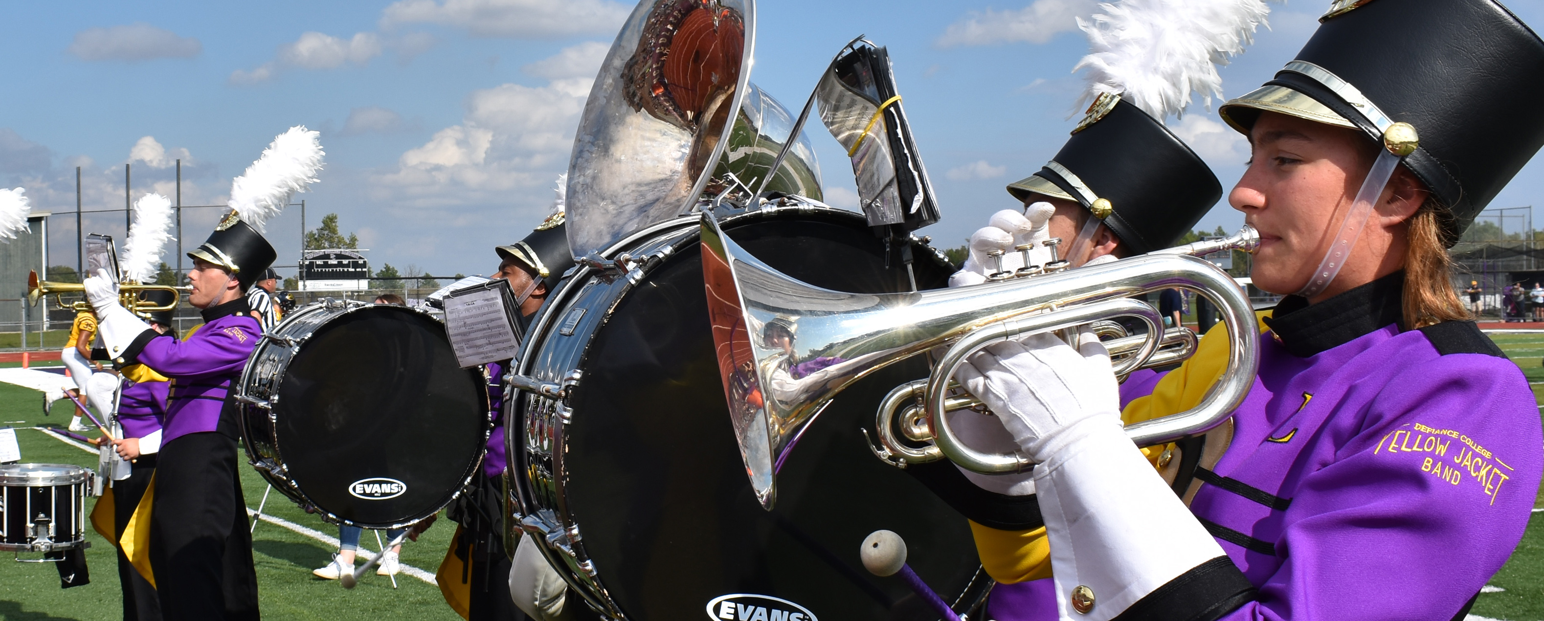 Four members of the Yellow Jacket Band playing their instruments on the football field in full uniforms of bright purple and gold.