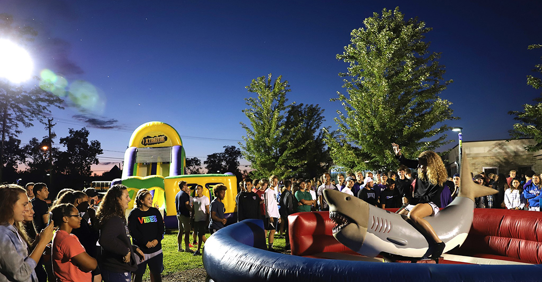 Large group of students gathered around an inflatable ring in which a student is riding on a spinning, mechanical shark.