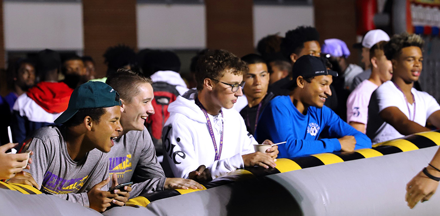 Five students leaning on an inflatable edge of an obstacle course, watching and laughing towards the middle of it off camera. Numerous students mingle in the background out of focus.