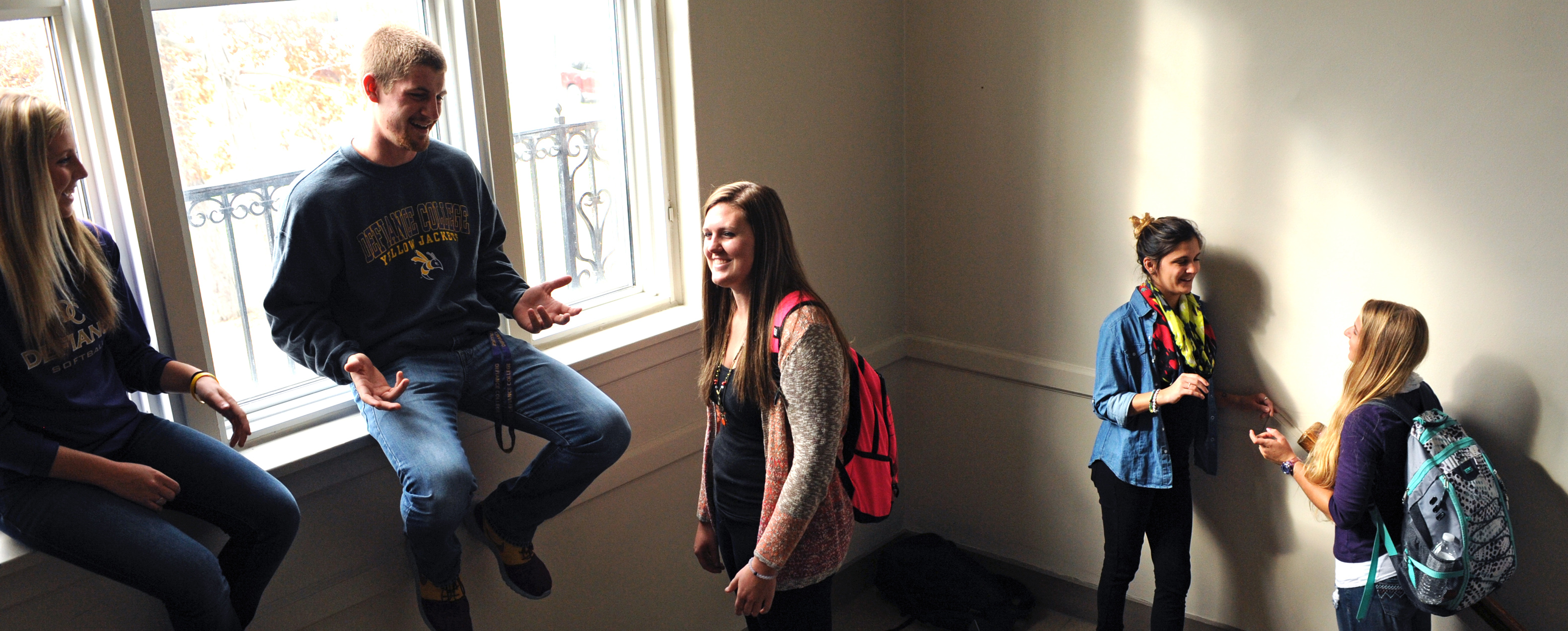 Five students gathered and speaking together in a large stairwell. Light from the big window in the background highlights the room.