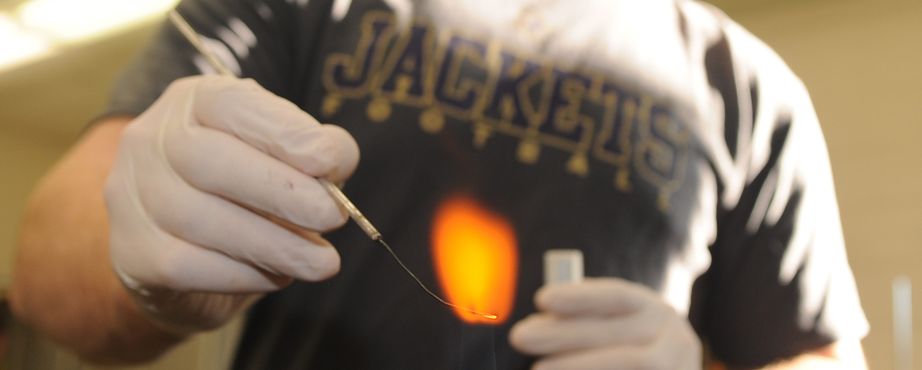 Close up of hands wearing rubber gloves heating a tool in a flame. Gloved hand in the background holding a test tube. Student's shirt is readable saying JACKETS Football.