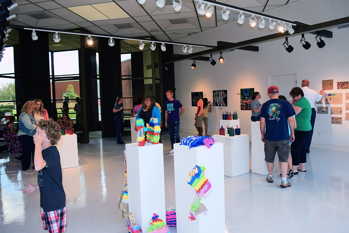 Multiple people in a white art gallery, looking at art on the walls and on white pedastals sitting around the middle of the room.