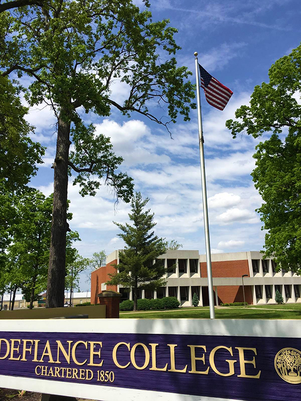 American flag flying against a blue sky in front of a Defiance College sign